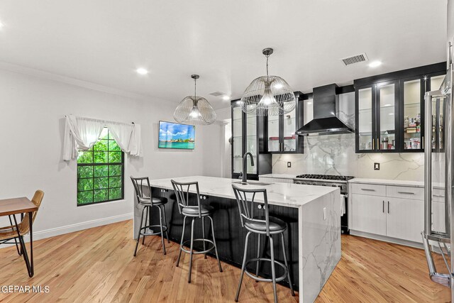 kitchen with sink, an island with sink, wall chimney exhaust hood, appliances with stainless steel finishes, and decorative light fixtures