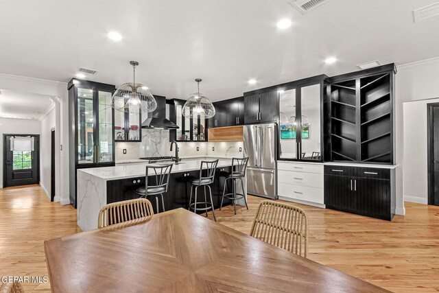 kitchen featuring stainless steel fridge, decorative light fixtures, a kitchen island with sink, wall chimney exhaust hood, and a kitchen bar