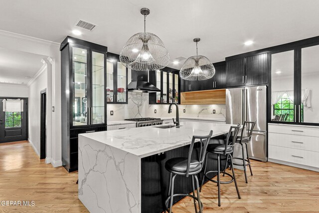 kitchen featuring light hardwood / wood-style floors, an island with sink, wall chimney range hood, appliances with stainless steel finishes, and decorative light fixtures