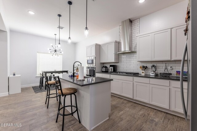 kitchen featuring light wood-type flooring, appliances with stainless steel finishes, a chandelier, wall chimney range hood, and a center island with sink