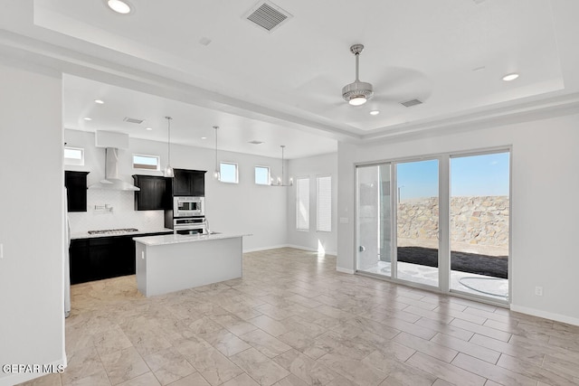 kitchen featuring a raised ceiling, backsplash, wall chimney exhaust hood, appliances with stainless steel finishes, and a center island with sink