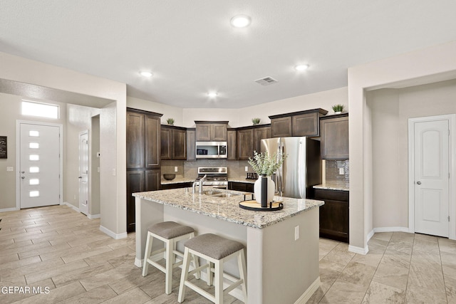 kitchen featuring appliances with stainless steel finishes, light stone countertops, a kitchen breakfast bar, decorative backsplash, and a kitchen island with sink