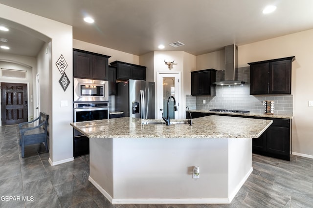 kitchen featuring tasteful backsplash, an island with sink, appliances with stainless steel finishes, wall chimney range hood, and a sink