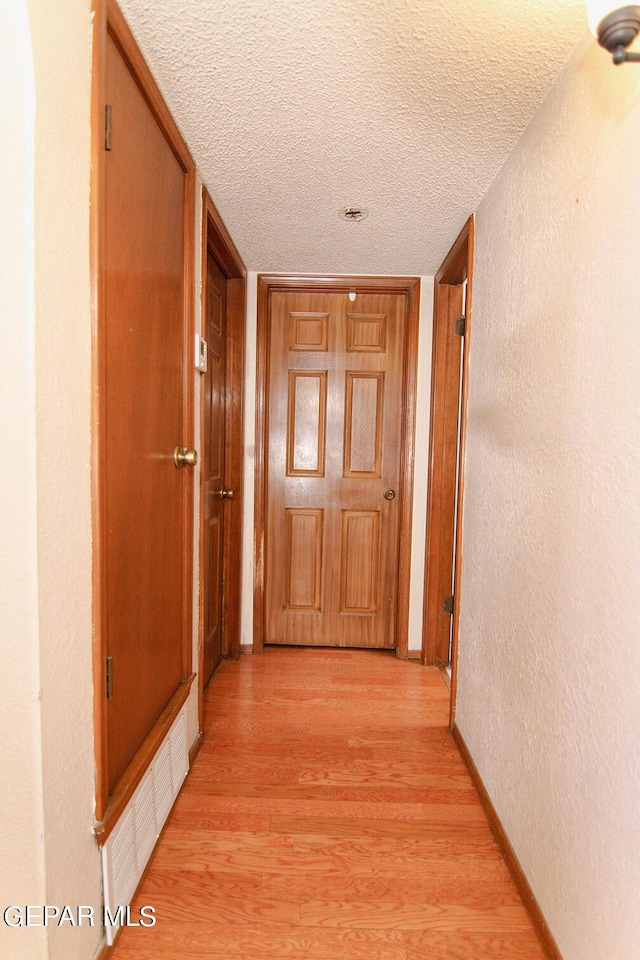 hall featuring a textured ceiling, a textured wall, visible vents, baseboards, and light wood finished floors