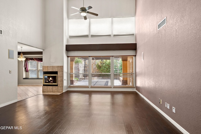 unfurnished living room with ceiling fan, a towering ceiling, hardwood / wood-style floors, and a tile fireplace