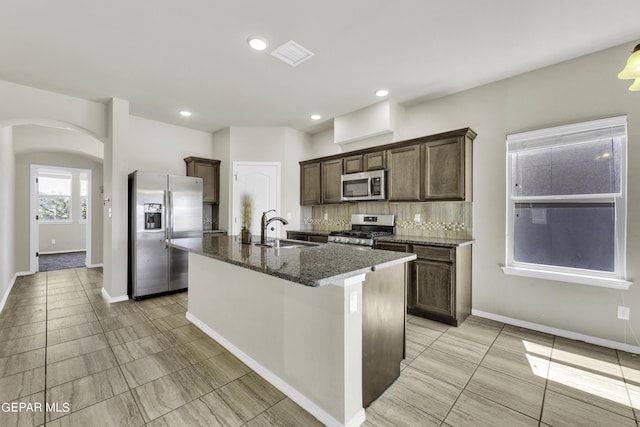 kitchen with dark stone countertops, arched walkways, a sink, dark brown cabinets, and appliances with stainless steel finishes