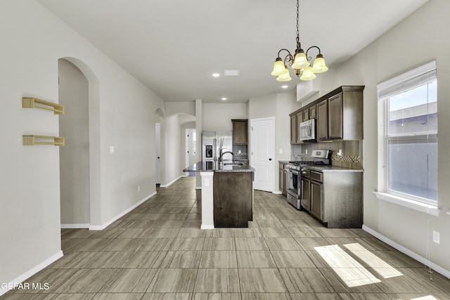 kitchen featuring a center island with sink, arched walkways, dark brown cabinets, appliances with stainless steel finishes, and a notable chandelier