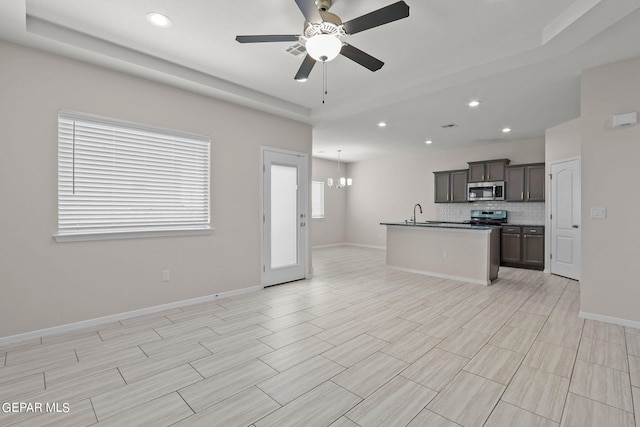 kitchen with a raised ceiling, stainless steel appliances, ceiling fan with notable chandelier, and a kitchen island with sink