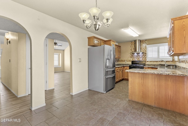 kitchen featuring black gas range oven, ceiling fan with notable chandelier, light wood-type flooring, stainless steel fridge with ice dispenser, and wall chimney range hood