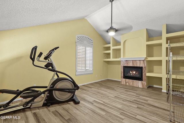 exercise room with light wood-type flooring, a textured ceiling, lofted ceiling, ceiling fan, and a premium fireplace