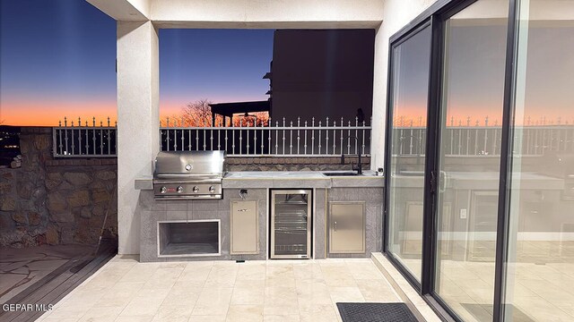 patio terrace at dusk with a grill, sink, a tile fireplace, and an outdoor kitchen