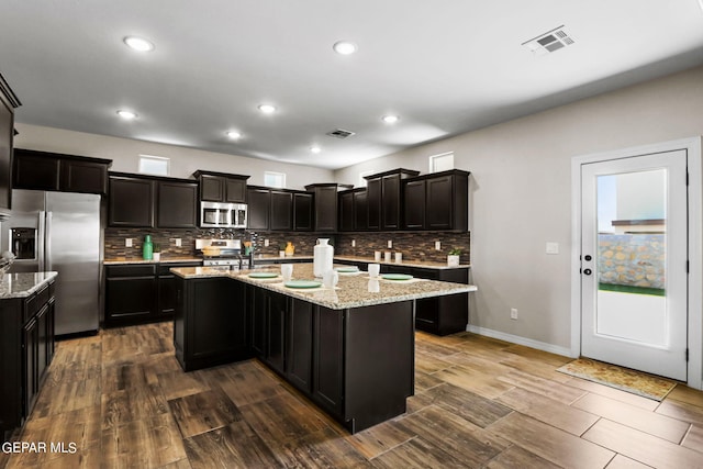 kitchen featuring appliances with stainless steel finishes, decorative backsplash, dark wood-type flooring, light stone counters, and a kitchen island with sink