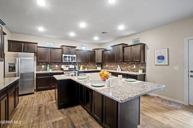 kitchen with dark brown cabinetry, wood-type flooring, a center island with sink, appliances with stainless steel finishes, and light stone countertops