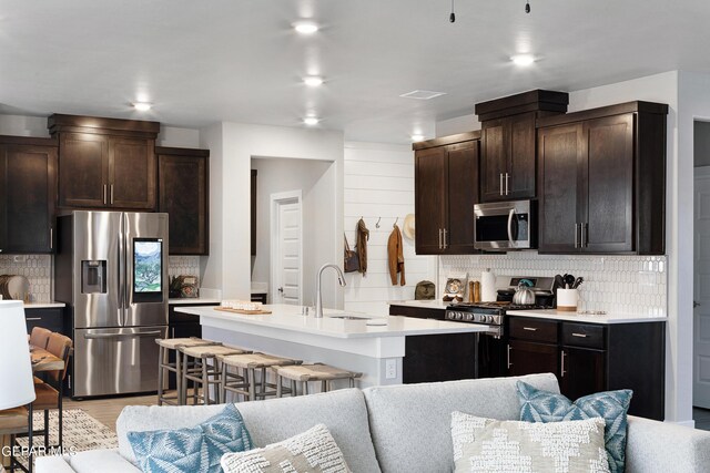 kitchen featuring a kitchen breakfast bar, an island with sink, tasteful backsplash, stainless steel appliances, and dark brown cabinetry