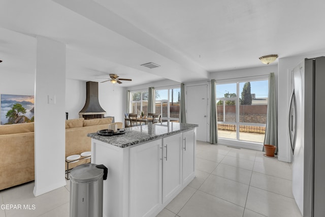 kitchen with an island with sink, white cabinets, stone counters, stainless steel refrigerator, and ceiling fan