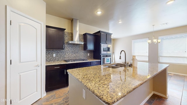 kitchen with sink, light stone counters, stainless steel appliances, a kitchen island with sink, and wall chimney range hood