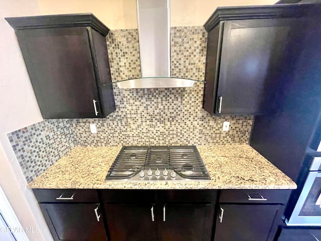 kitchen with backsplash, dark brown cabinets, wall chimney exhaust hood, light stone counters, and stainless steel gas stovetop