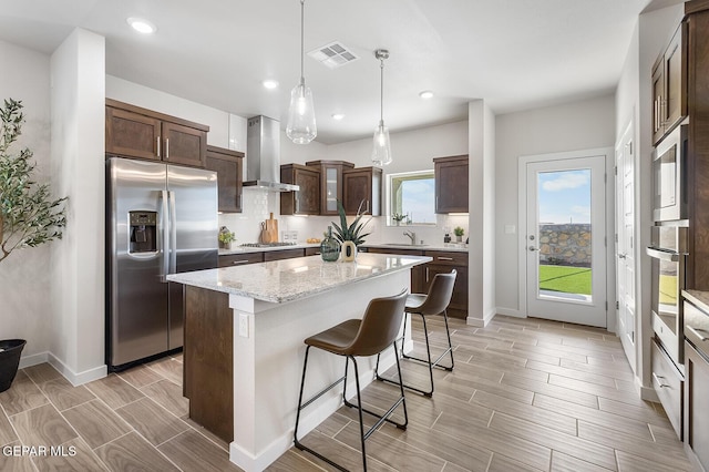 kitchen featuring stainless steel appliances, a kitchen island, visible vents, hanging light fixtures, and wall chimney range hood