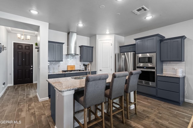 kitchen with a breakfast bar, wall chimney exhaust hood, stainless steel appliances, a center island with sink, and light stone countertops