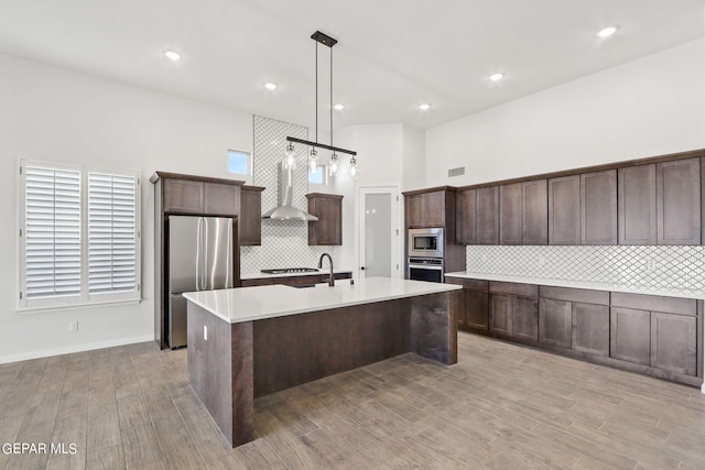 kitchen with light hardwood / wood-style floors, stainless steel appliances, decorative light fixtures, a center island with sink, and wall chimney range hood