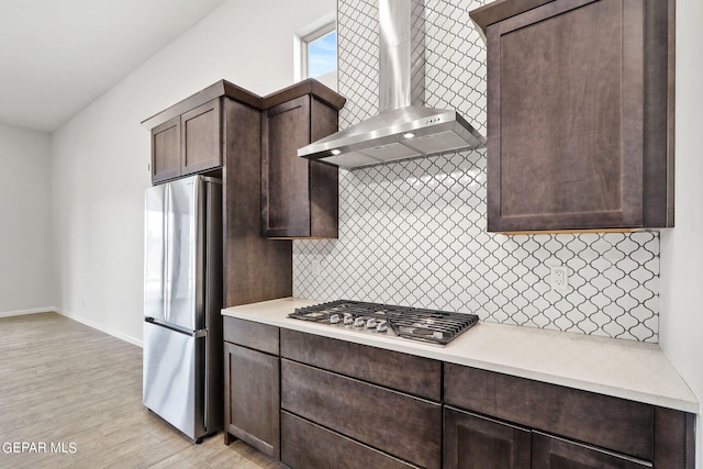 kitchen featuring dark brown cabinets, light wood-type flooring, tasteful backsplash, wall chimney exhaust hood, and appliances with stainless steel finishes