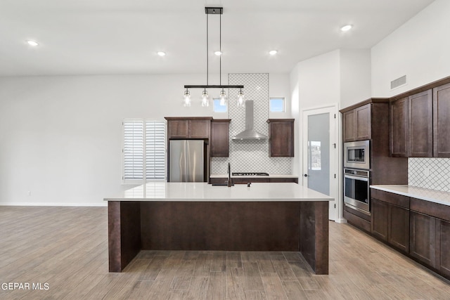 kitchen with pendant lighting, light hardwood / wood-style floors, a kitchen island with sink, wall chimney range hood, and stainless steel appliances