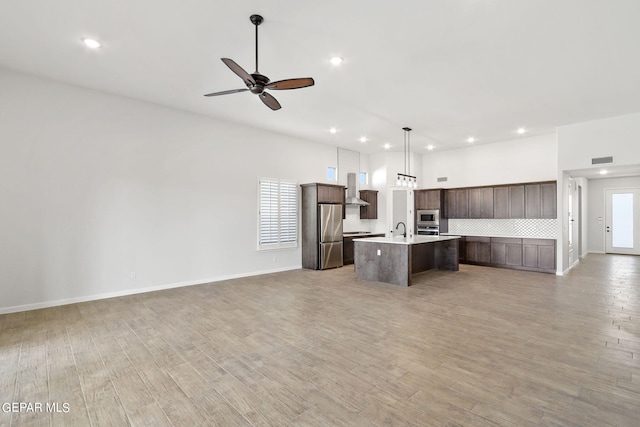 kitchen with pendant lighting, a center island with sink, wall chimney exhaust hood, appliances with stainless steel finishes, and light hardwood / wood-style floors