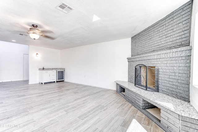 unfurnished living room with ceiling fan, wine cooler, a textured ceiling, light hardwood / wood-style flooring, and a fireplace