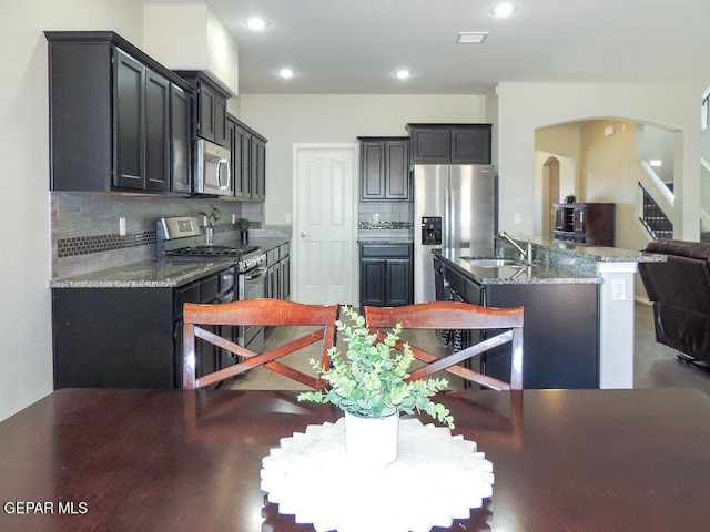 kitchen with stainless steel appliances, backsplash, a center island with sink, sink, and stone counters