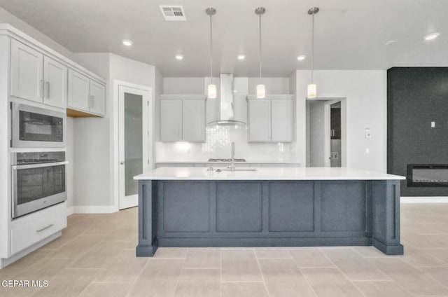 kitchen featuring a center island with sink, wall chimney exhaust hood, hanging light fixtures, and stainless steel oven