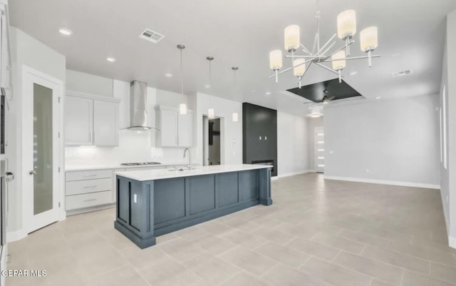 kitchen with white cabinets, a center island with sink, wall chimney exhaust hood, decorative light fixtures, and an inviting chandelier