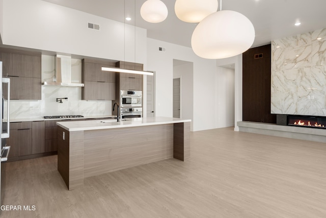 kitchen with a kitchen island with sink, wall chimney range hood, hanging light fixtures, a premium fireplace, and light hardwood / wood-style floors