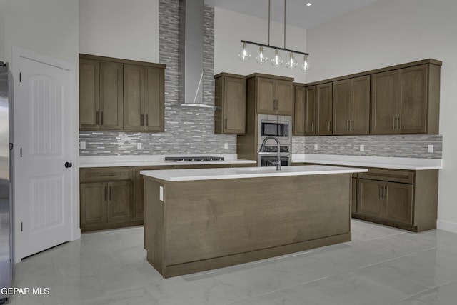 kitchen featuring a center island with sink, decorative light fixtures, a high ceiling, appliances with stainless steel finishes, and wall chimney exhaust hood