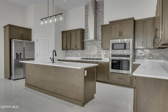 kitchen featuring sink, a towering ceiling, wall chimney range hood, stainless steel appliances, and an island with sink