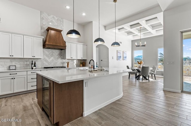 kitchen featuring coffered ceiling, white cabinets, a center island with sink, decorative light fixtures, and custom range hood