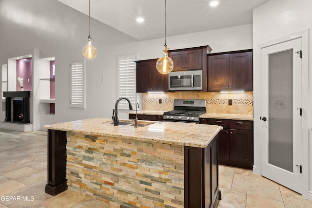 kitchen with an island with sink, appliances with stainless steel finishes, light stone countertops, pendant lighting, and sink