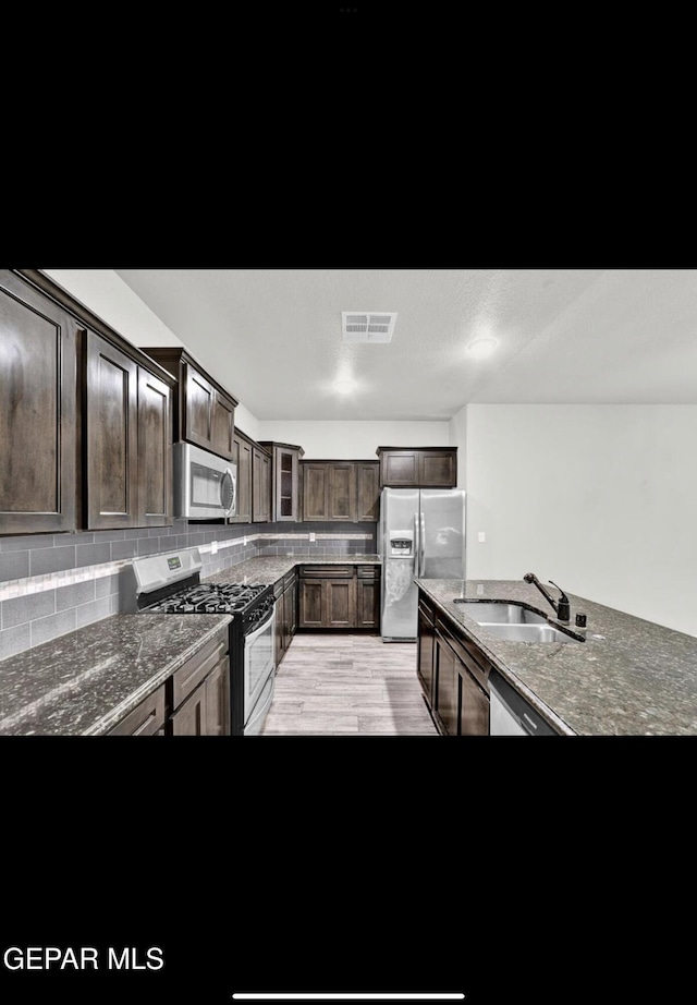 kitchen with appliances with stainless steel finishes, light hardwood / wood-style flooring, dark stone countertops, dark brown cabinetry, and sink