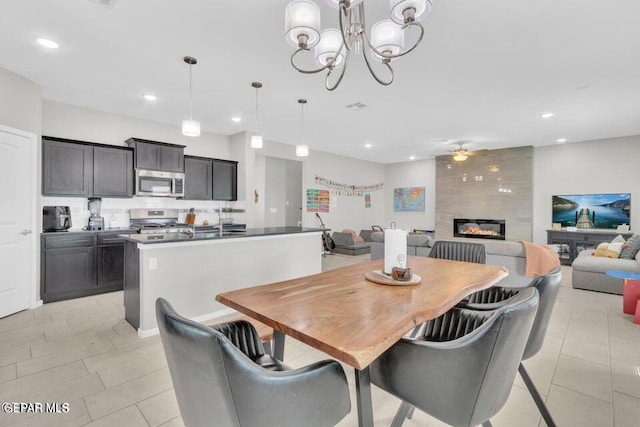 dining area with light tile patterned flooring, ceiling fan with notable chandelier, and a large fireplace