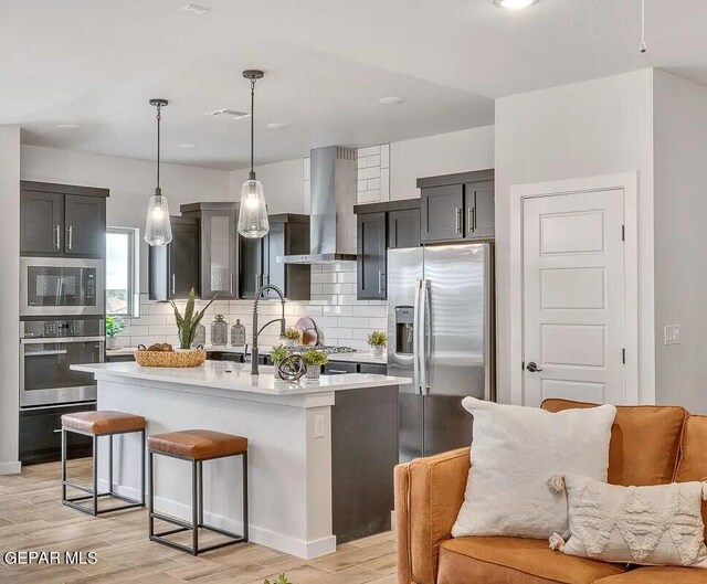 kitchen with hanging light fixtures, a center island with sink, light hardwood / wood-style floors, wall chimney exhaust hood, and stainless steel appliances