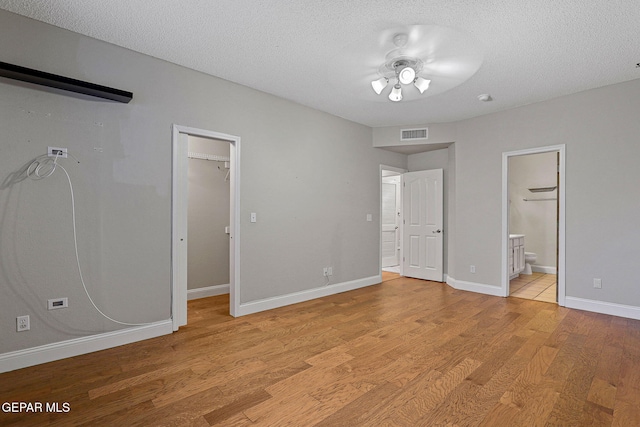 unfurnished bedroom with ceiling fan, a textured ceiling, a spacious closet, connected bathroom, and light wood-type flooring