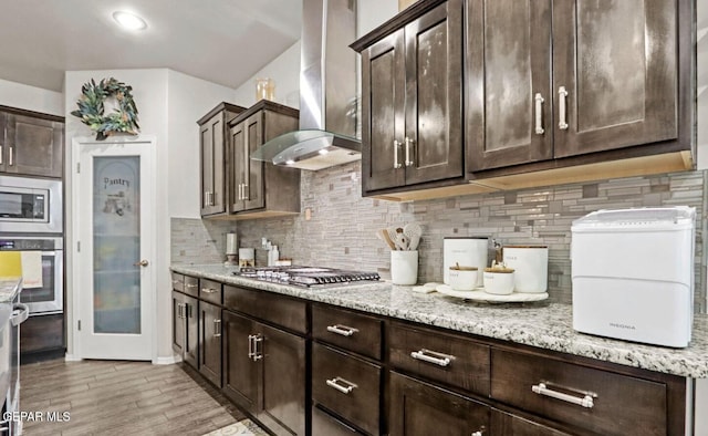 kitchen featuring dark brown cabinetry, wall chimney range hood, light stone counters, backsplash, and appliances with stainless steel finishes