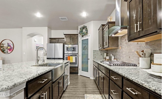 kitchen featuring backsplash, wall chimney range hood, light stone countertops, dark brown cabinets, and stainless steel appliances