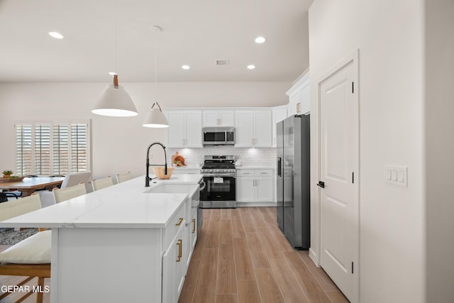 kitchen featuring hanging light fixtures, appliances with stainless steel finishes, white cabinetry, a kitchen island with sink, and light wood-type flooring