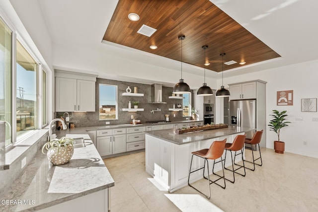 kitchen with wall chimney exhaust hood, stainless steel appliances, backsplash, pendant lighting, and a kitchen island