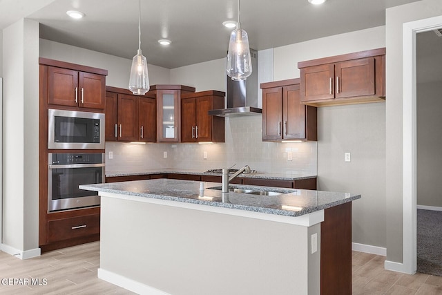 kitchen with tasteful backsplash, wall chimney range hood, light wood-type flooring, an island with sink, and stainless steel appliances