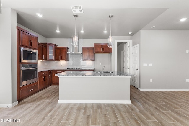 kitchen with wall chimney exhaust hood, appliances with stainless steel finishes, light wood-type flooring, and a kitchen island with sink