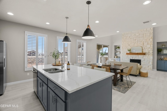 kitchen with hanging light fixtures, a kitchen island with sink, light wood-type flooring, a stone fireplace, and sink