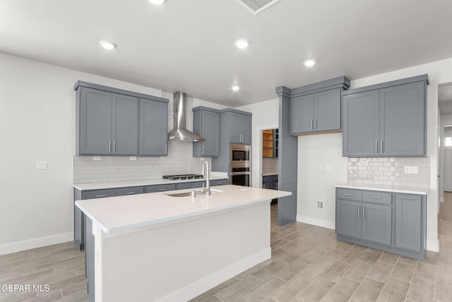 kitchen featuring sink, appliances with stainless steel finishes, a kitchen island with sink, wall chimney range hood, and gray cabinetry