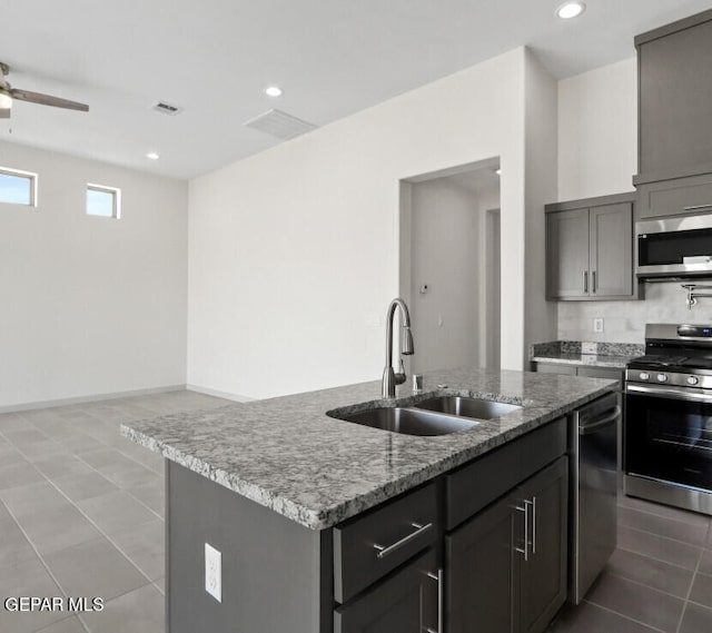 kitchen featuring stainless steel appliances, a center island with sink, sink, light stone counters, and ceiling fan