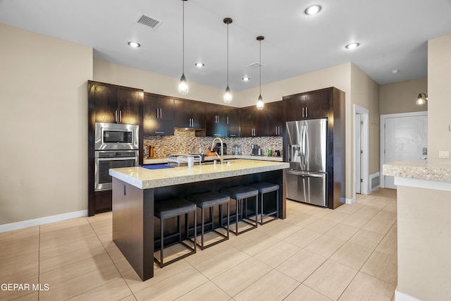 kitchen featuring dark brown cabinetry, appliances with stainless steel finishes, tasteful backsplash, an island with sink, and pendant lighting